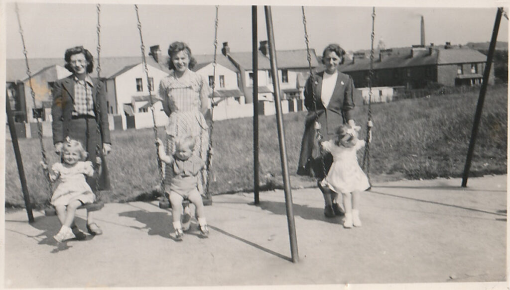 3 mothers standing behind their children who are on the swings in the playground, 1951